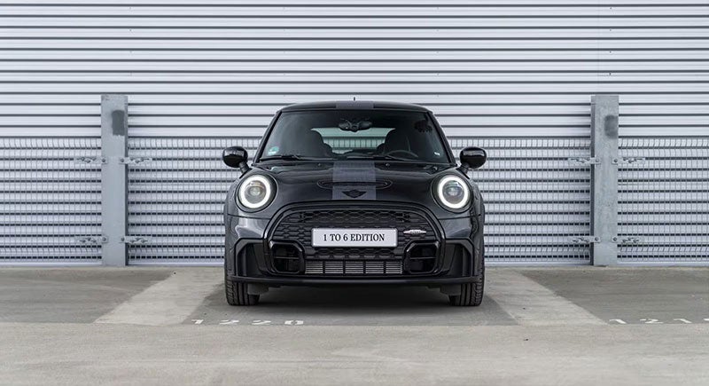 Front view of a MINI John Cooper Works 1to6 Edition in Midnight Black II body color, parked on a pavement surface with a metal wall and metal fencing behind it.