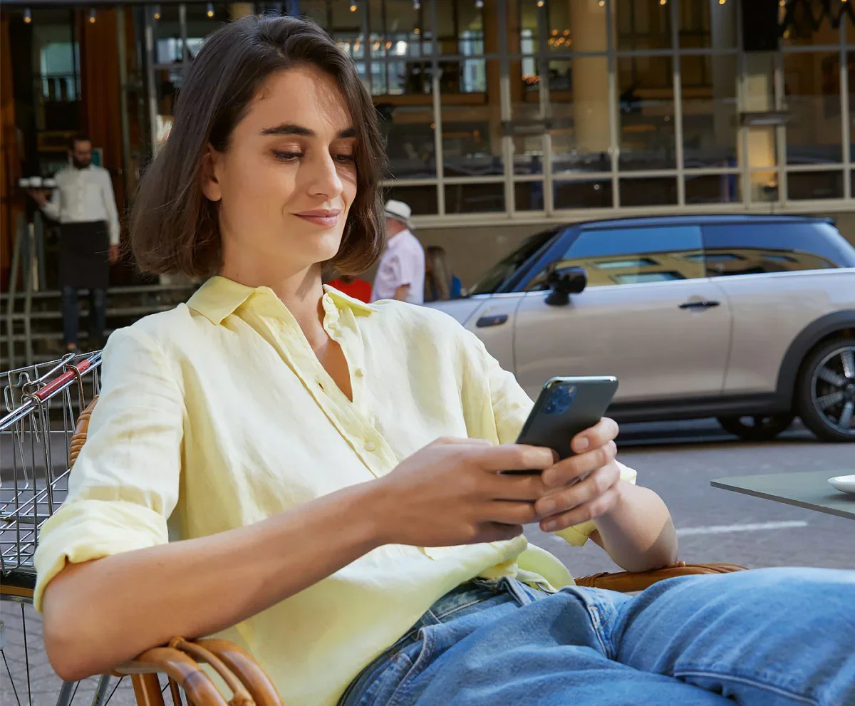 Female wearing yellow collared button-down shirt and jeans, sitting in a chair and holding a smartphone next to a street with a grey MINI vehicle parked in the background. | Tom Bush MINI in Jacksonville FL
